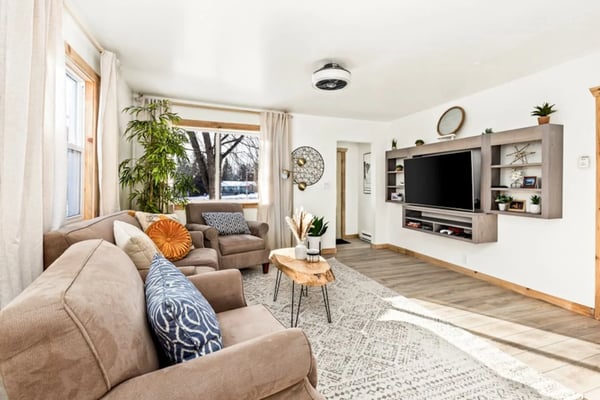 Modern living room with stone fireplace, wood beams, multiple windows with mountain views, and comfortable seating around wooden coffee table