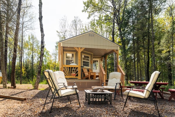 Modern glamping cabin with covered porch, wooden Adirondack chairs, and deck surrounded by forest trees.
